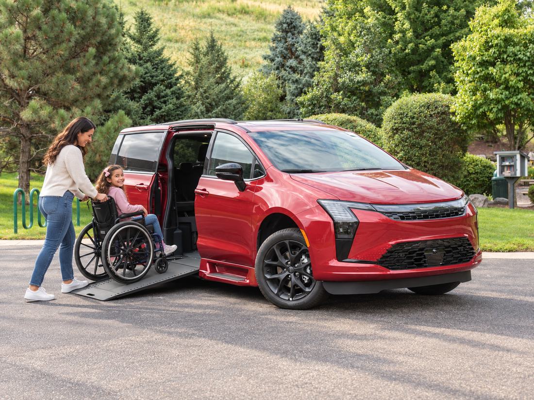 A mother and her young daughter, who uses a wheelchair, at the deployed side-entry ramp of a red Chrysler Pacifica on a neighbourhood street